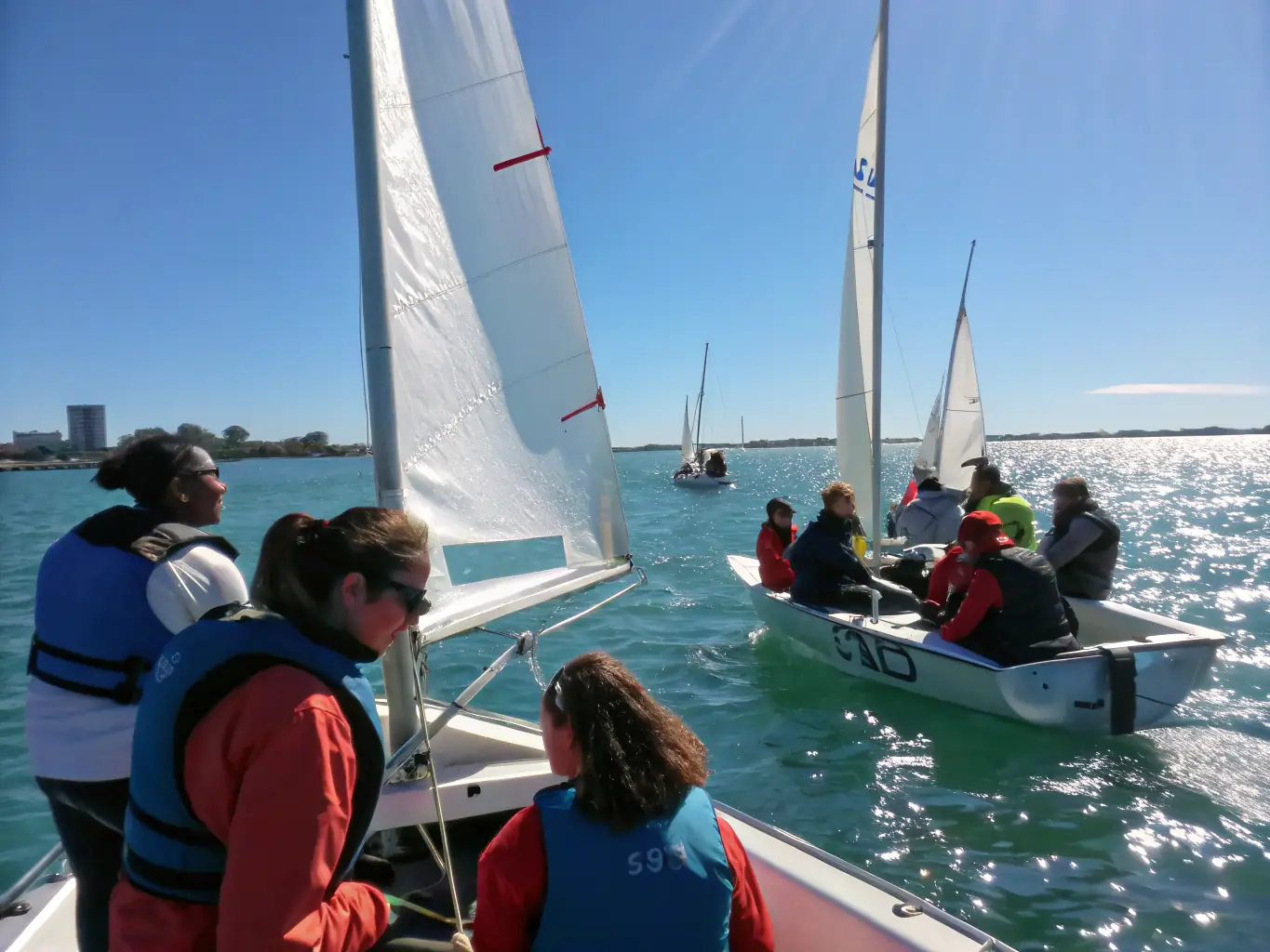 A group of business professionals are laughing and working together to raise the sail on a catamaran during an Achilles Challenge event, showcasing teamwork and collaboration.